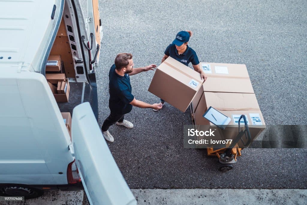 Two delivery workers transferring cardboard boxes from a white van onto a yellow pallet jack.