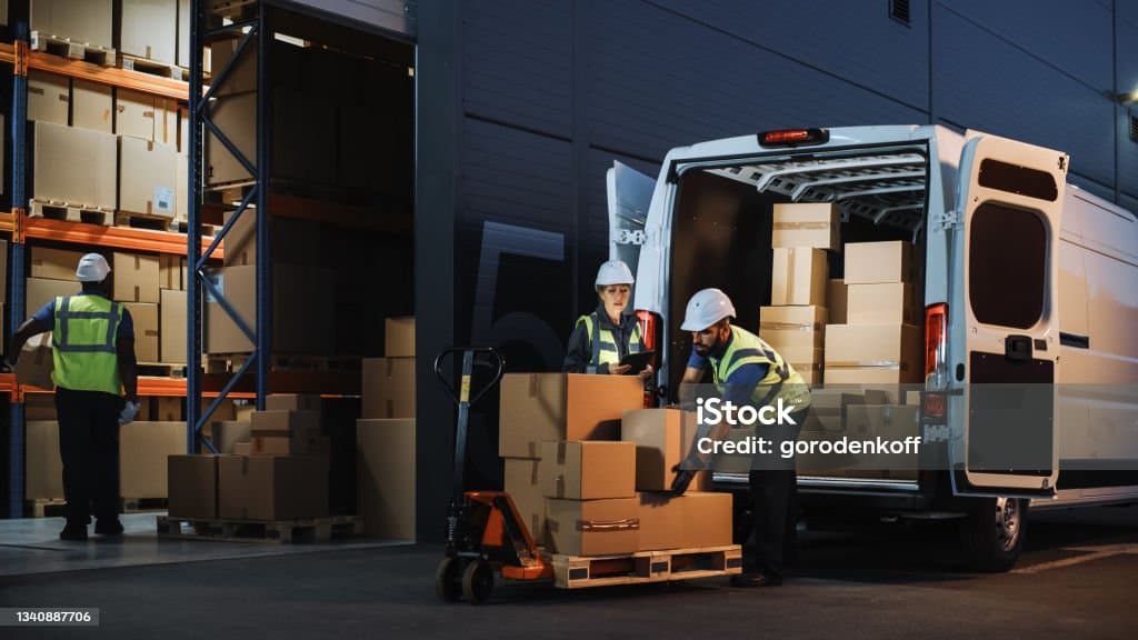 Logistics workers in safety vests loading cardboard boxes into a white delivery van at night.